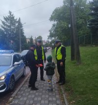 Dwaj umundurowani funkcjonariusze ruchu drogowego stojąc przy oznakowanym radiowozie wręczają kamizelkę odblaskową mężczyźnie, który wyszedł ze szkoły ze swoim dzieckiem