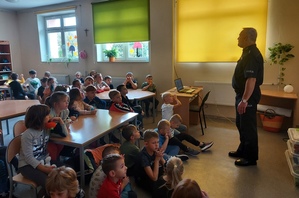 umundurowany policjant prowadzi prelekcję dla dzieci ze Szkoły Podstawowej nr 3 w Brzezinach