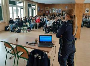 umundurowany policjant prowadzi prelekcję dla uczniów, który na siedząco słuchają wykładu.