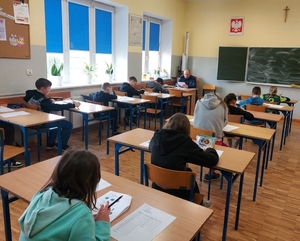 uczniowie siedząc w szkolnych ławkach piszą test.