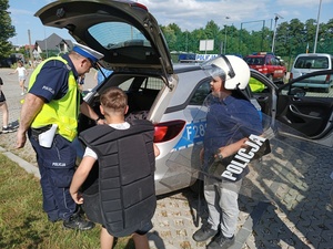 dzieci przy oznakowanym policyjnym radiowozie przymierzają strój chroniący policjanta podczas zabezpieczania imprez masowych.
