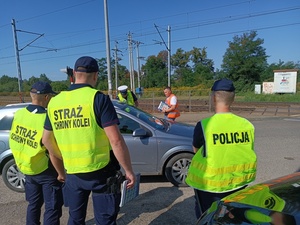 umundurowany policjant wręcza ulotki kierowcy samochodu stojącego przed przejazdem kolejowym, a obok stoi kolejny policjant i służba ochrony kolei.