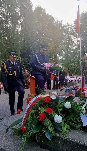 umundurowany policjant wraz z umundurowanym strażakiem składają kwiaty pod pomnikiem poległych żołnierzy.