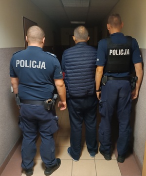 dwaj umundurowani policjanci prowadzą korytarzem zatrzymanego mężczyznę.