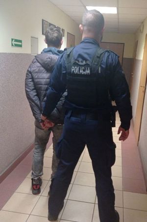 umundurowany policjant w kamizelce z napisem policja prowadzi korytarzem zatrzymanego mężczyznę.