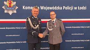 Na tle baneru Komendy Wojewódzkiej Policji w Łodzi Komendant inspektor Marcin Grzelak gratuluje aspirantowi sztabowemu Przemysławowi Goli podając mu rękę.