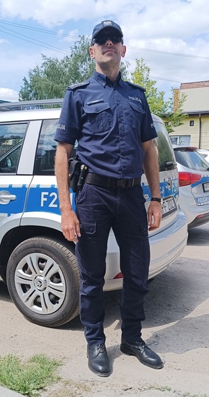 aspirat sztabowy Grzegorz Miazek stojący w mundurze przy oznakowanym policyjnym radiowozie.