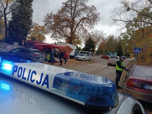 na dachu oznakowanego radiowozu napis policja, a w tle umundurowany policjant kontroluje samochód.