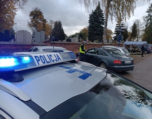 na dachu oznakowanego radiowozu napis policja, a w tle umundurowany policjant kontroluje samochód.