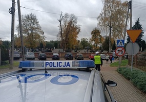 na dachu oznakowanego radiowozu napis policja, a w tle umundurowany policjant kontroluje samochód.