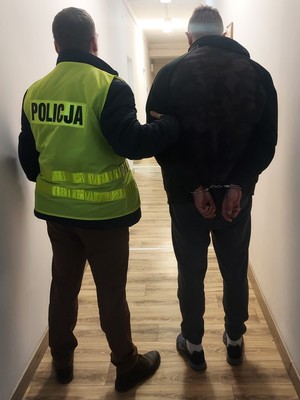 nieumundurowany policjant z kamizelką z napisem Policja prowadzi korytarzem zatrzymanego mężczyznę.