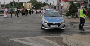 umundurowany policjant stojąc przy oznakowanym radiowozie zabezpiecza przejście procesji.
