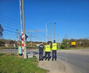 umundurowani policjanci strażnicy ochrony kolei stoją przy otwartym przejeździe kolejowym.