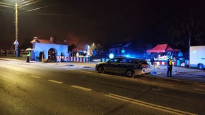 umundurowani policjanci stoją przy oznakowanym radiowozie i pilnują porządku przy cmentarnym wejściu.