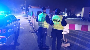 umundurowani policjanci stoją przy oznakowanym radiowozie i pilnują porządku przy cmentarnym wejściu.