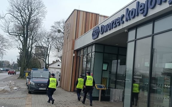Umundurowany policjant wraz z funkcjonariuszami Służby Ochrony Kolei przed budynkiem PKP .