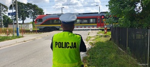 Policjant kontroluje przejazd kolejowy, przez który przejeżdża pociąg osobowy.